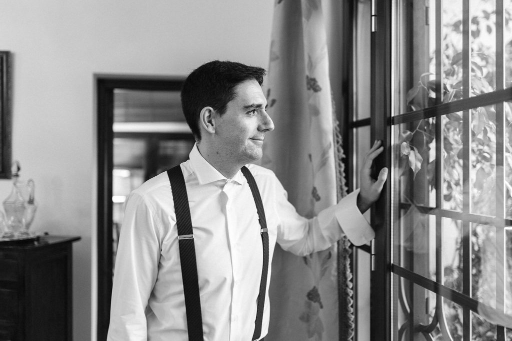 Novio ajustando los tirantes junto a una ventana con reja, momento previo a la ceremonia