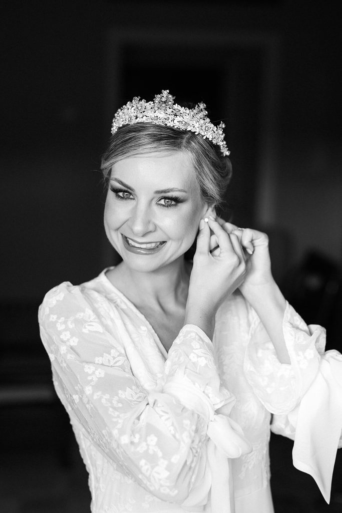 Novia sonriente ajustándose un pendiente, vestida con bata de encaje blanco